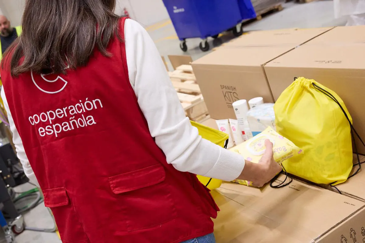 Una mujer de espaldas, con chaleco rojo con el rótulo «cooperación española», coloca productos de higiene y un paquete de toallitas junto a una bolsa amarilla sobre grandes cajas de cartón en una nave logística. Sobre las cajas se lee «Spanish Kits». Al fondo, un contenedor azul y palés de madera.
