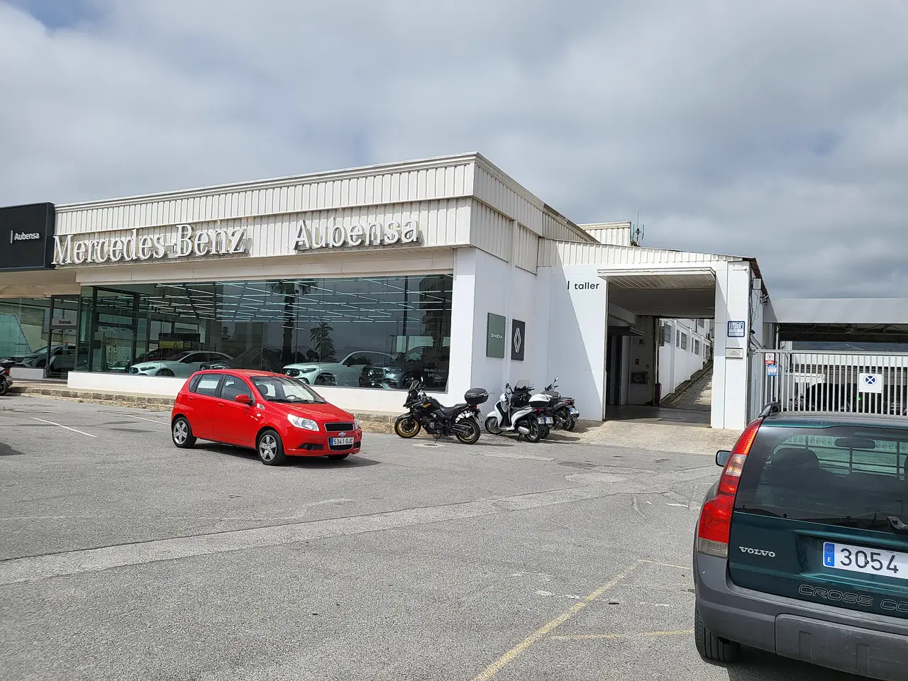 Nave blanca de un concesionario de automóviles con los rótulos «Mercedes-Benz» y «Aubensa» sobre el escaparate, coches nuevos tras el cristal, un coche rojo y varias motos aparcados delante y la entrada a un taller a la derecha, en una explanada de asfalto bajo un cielo nublado.