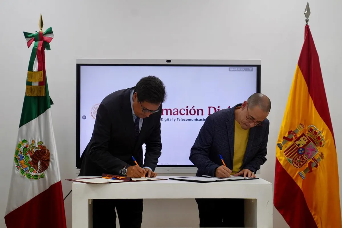 Dos hombres firman sendos documentos sobre una mesa blanca, entre una bandera de México y una de España, ante una pantalla con el logotipo de la Agencia de Transformación Digital y Telecomunicaciones.