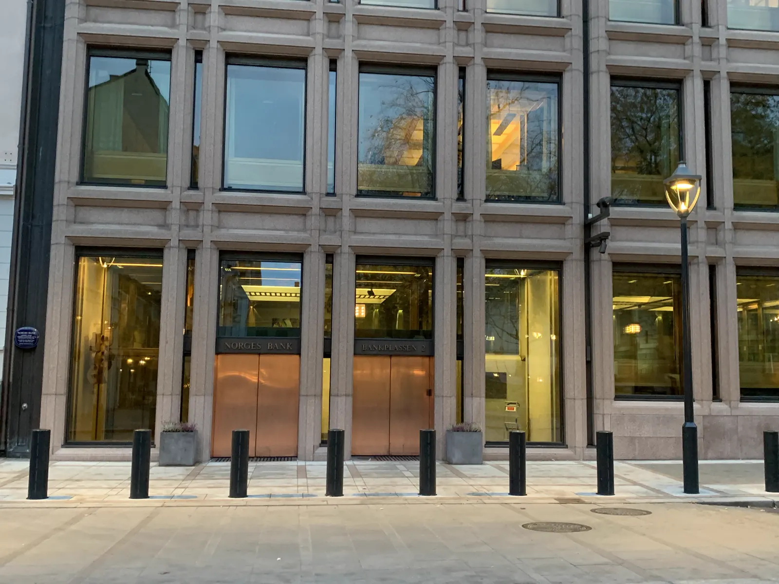 Entrada del edificio de Norges Bank en Bankplassen 2 de Oslo, con puertas de cobre bajo la inscripción «Norges Bank» y bolardos negros en la acera.