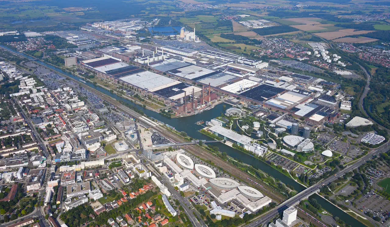 Vista aérea de la fábrica de Volkswagen en Wolfsburgo: una hilera de naves industriales de cubierta blanca junto al canal, con las cuatro chimeneas de ladrillo de la central térmica y, al otro lado del agua, los pabellones y torres de cristal del parque Autostadt.