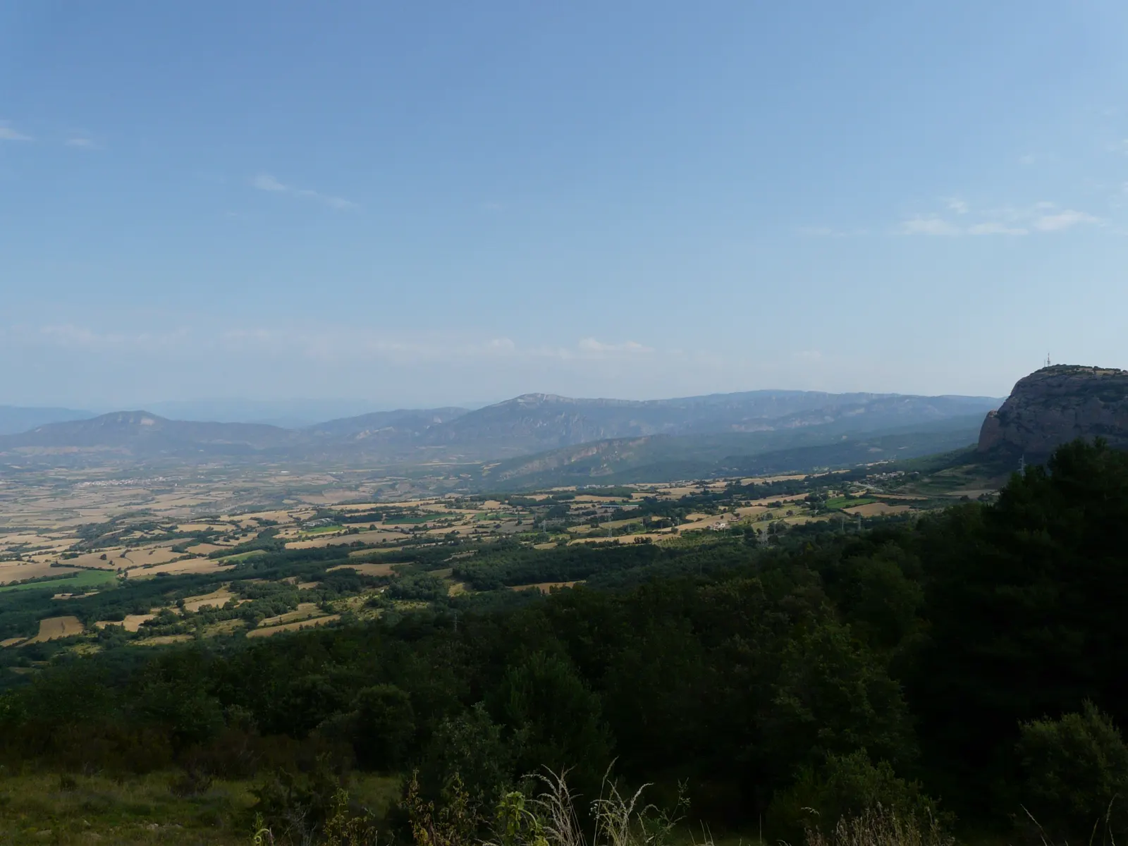 Vista de la Conca de Tremp desde una loma cubierta de matorral y pinos: al fondo, un mosaico llano de parcelas de rastrojo amarillo y campos verdes se extiende hasta una sierra alargada de roca clara; a la derecha, un farallón rocoso con una antena en la cima cierra el valle bajo un cielo azul con calima.