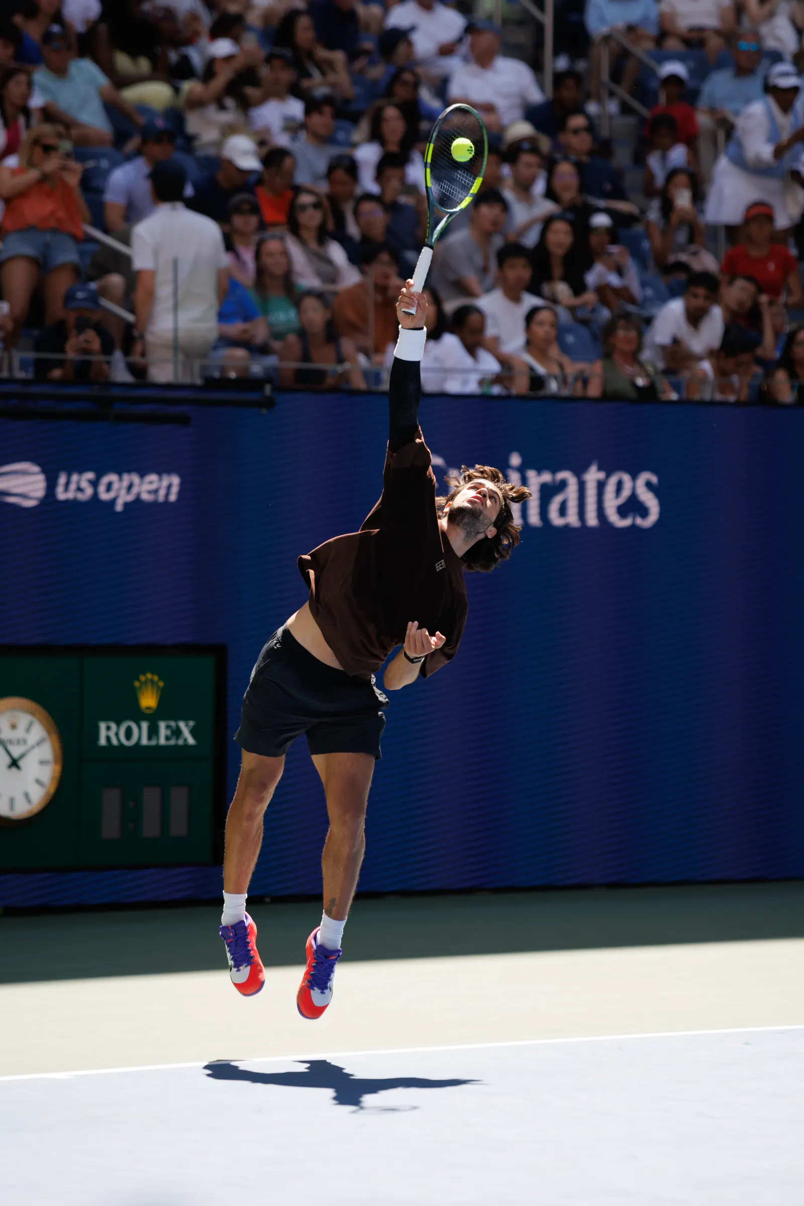 Un tenista de camiseta oscura salta con el brazo derecho estirado para golpear un saque; la pelota está aún sobre el cordaje. Al fondo, el panel azul de la pista con el rótulo «us open», un reloj y la grada llena.
