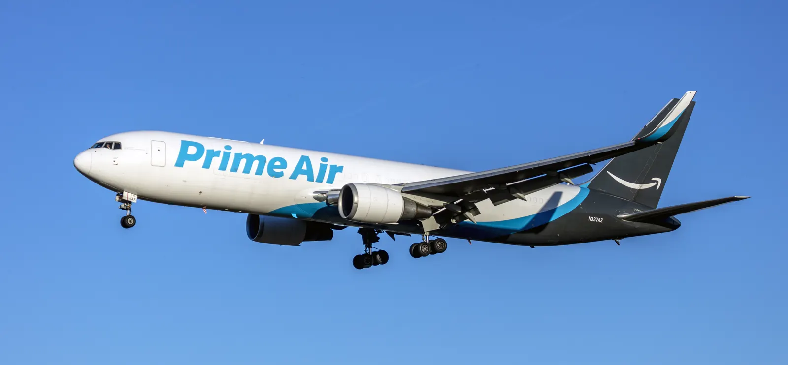 Un Boeing 767 de carga blanco con el rótulo azul Prime Air en el fuselaje y la sonrisa de Amazon en la cola, volando con el tren de aterrizaje desplegado contra un cielo despejado