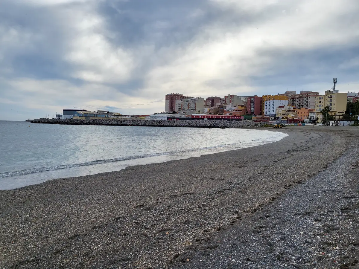 Playa de arena y grava sin bañistas, con la orilla en calma a la izquierda, un espigón de escollera al fondo y, tras él, naves industriales y bloques de viviendas de colores de Ceuta bajo un cielo nublado.
