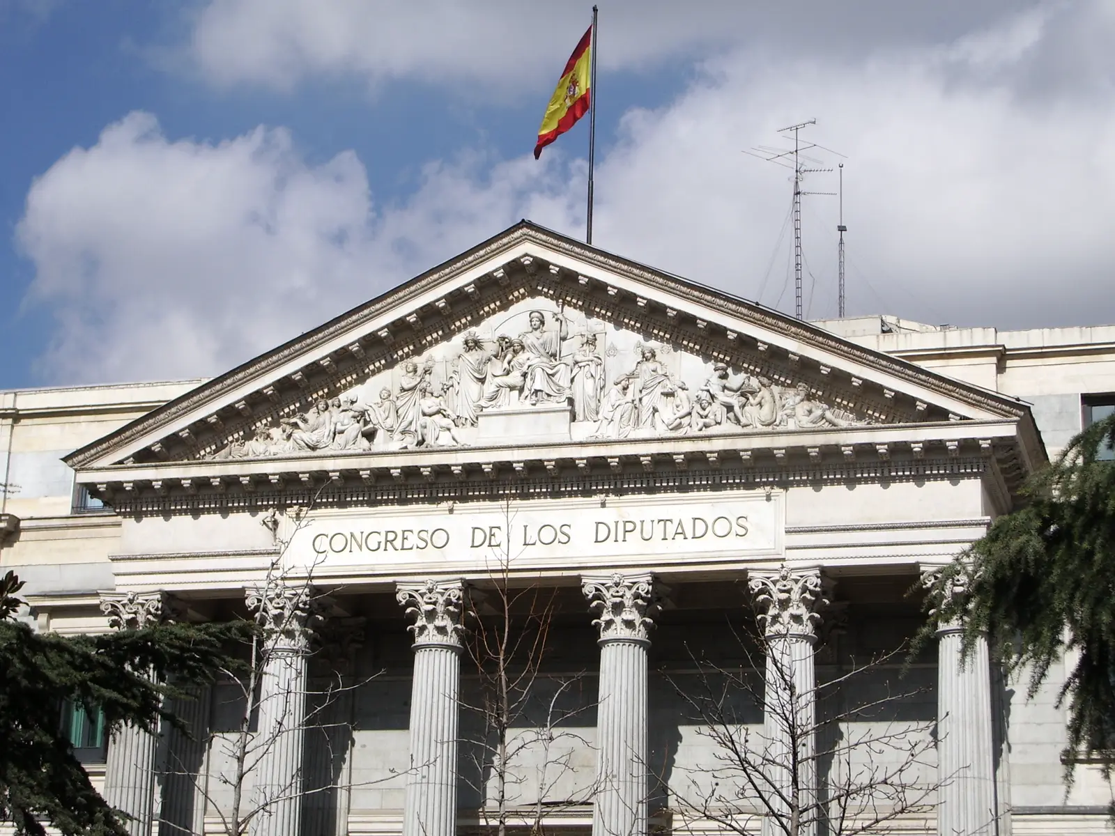 Fachada principal del Congreso de los Diputados: frontón triangular con relieve escultórico, la inscripción «CONGRESO DE LOS DIPUTADOS» y seis columnas corintias, con la bandera de España ondeando sobre el tejado y ramas de árbol en primer plano.