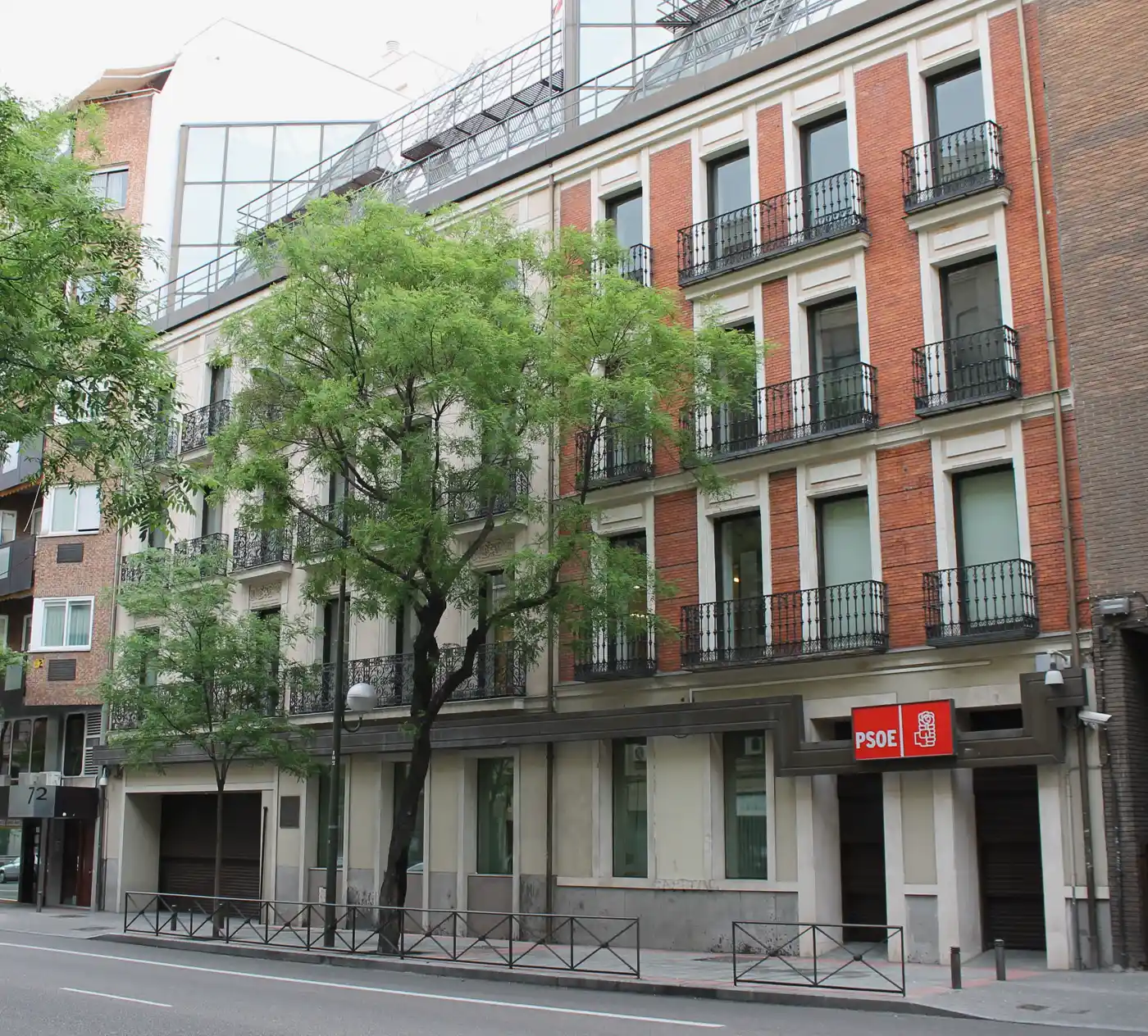 Fachada de la sede federal del PSOE en la calle Ferraz de Madrid: un edificio de cuatro plantas de ladrillo rojo y piedra clara, con balcones de reja negra y un cartel rojo con el rótulo «PSOE» y el logotipo del puño y la rosa sobre la entrada; delante, un árbol y la calle vacía.