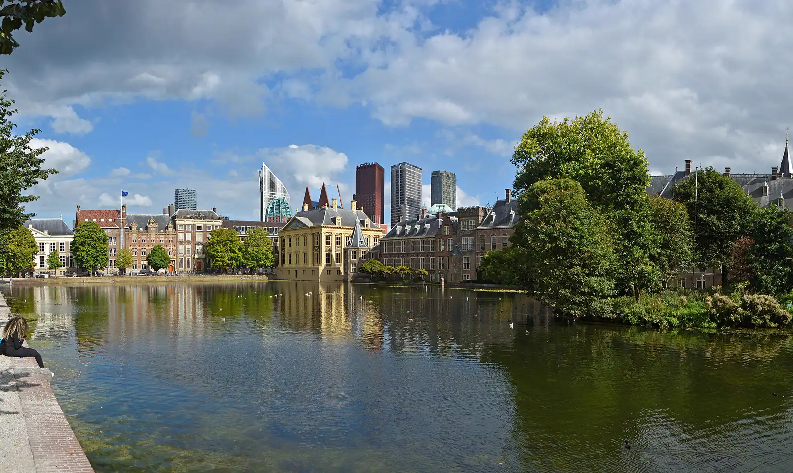 Estanque Hofvijver de La Haya con los edificios del Binnenhof y el museo Mauritshuis al fondo, una bandera de la Unión Europea a la izquierda y las torres de oficinas de la ciudad en el horizonte.