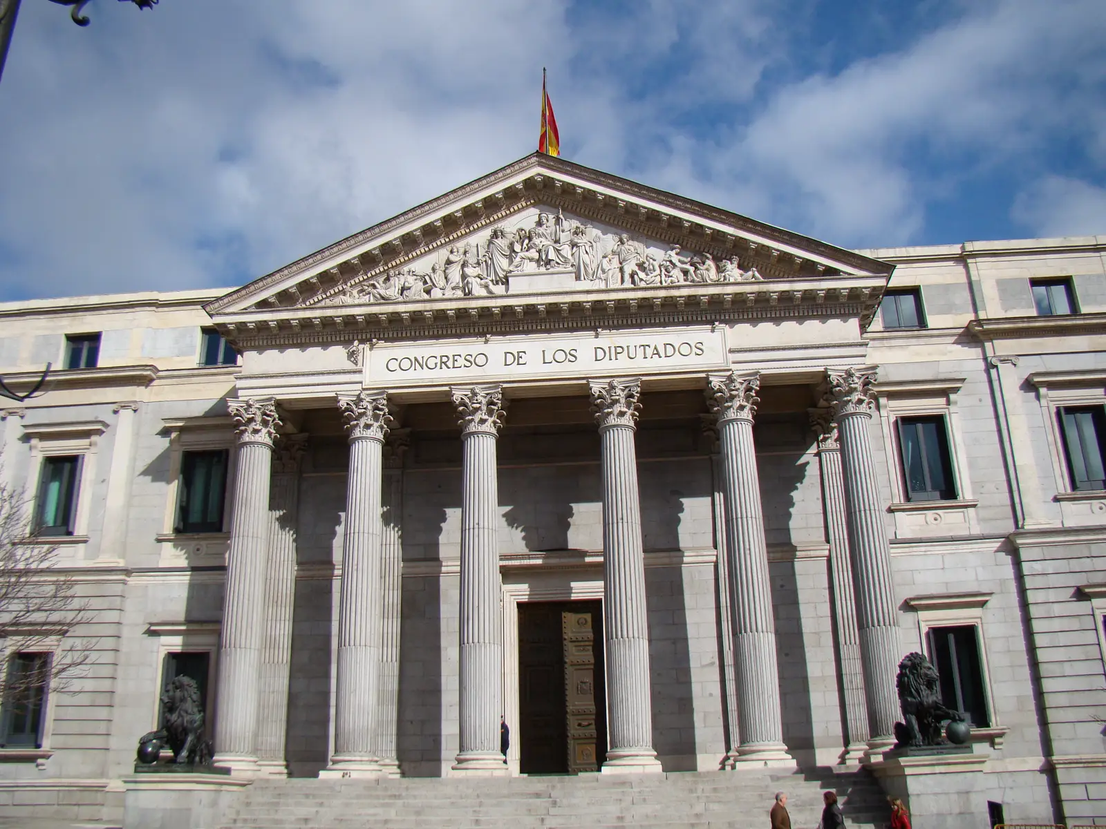 Fachada principal del Palacio de las Cortes, sede del Congreso de los Diputados: seis columnas corintias, frontón con relieve escultórico, la inscripción «CONGRESO DE LOS DIPUTADOS», los dos leones de bronce a los lados de la escalinata y la bandera de España sobre el tejado, bajo un cielo azul con nubes.