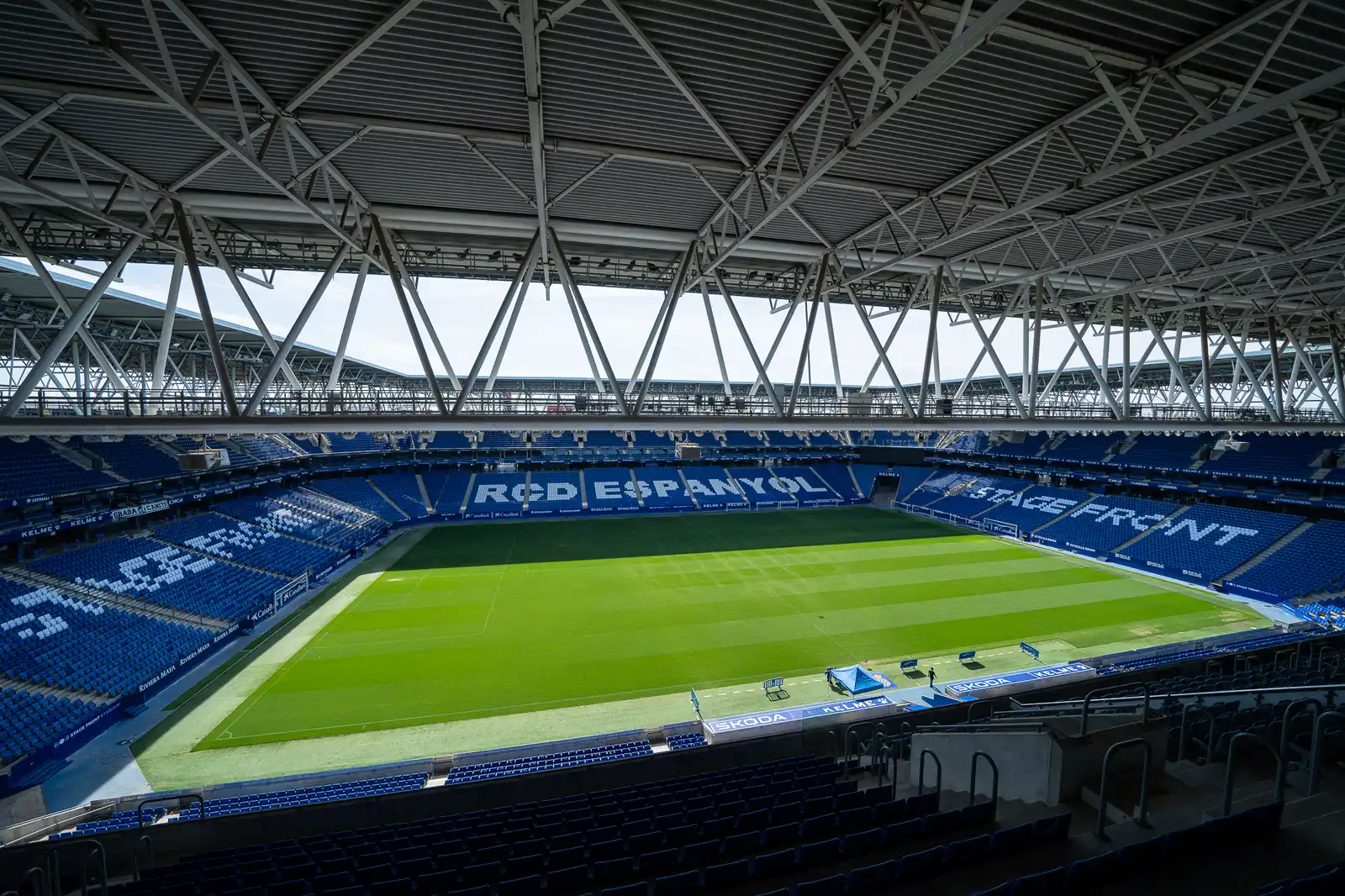 Interior vacío del estadio del RCD Espanyol en Cornellà, con el césped rayado, la cubierta metálica blanca y las gradas azules en las que se lee «RCD Espanyol» y «Stage Front» formado con asientos blancos.