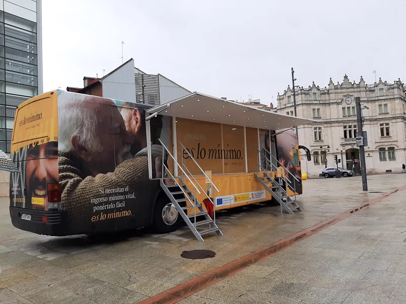 Autobús rotulado con el lema «es lo mínimo» y con fotografías de personas mayores, aparcado y abierto al público con una marquesina y dos escaleras de acceso en una plaza empedrada.