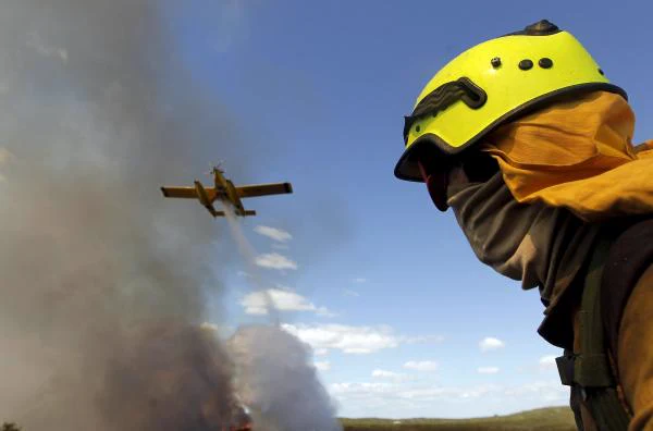 Un brigadista forestal de espaldas, en primer plano a la derecha, con casco amarillo flúor, gafas y capucha ignífuga naranja cubriéndole el rostro, mira hacia la izquierda del encuadre. Allí, sobre una columna de humo gris que sale de un frente de llamas anaranjado a ras de suelo, un pequeño avión amarillo de un solo motor y con flotadores suelta un chorro de agua. Cielo azul con nubes bajas al fondo.