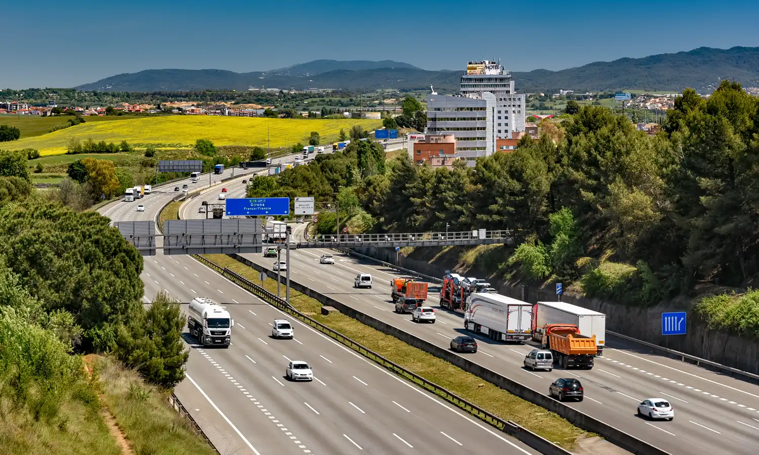 Vista elevada de una autopista de varios carriles con turismos, furgonetas y camiones circulando en ambos sentidos; junto a la calzada, un cartel azul indica la dirección a Girona y Francia, y al fondo se ven campos amarillos, un edificio de oficinas y colinas boscosas.