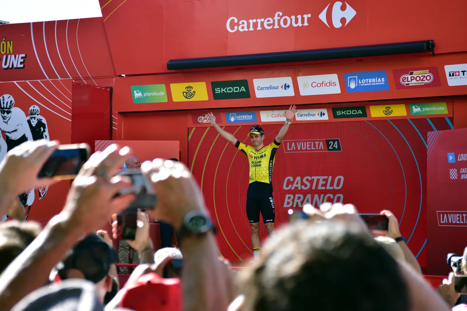 Un ciclista con el maillot amarillo y negro del Visma-Lease a Bike levanta los dos brazos sobre un podio rojo de La Vuelta; delante, decenas de manos del público lo graban con teléfonos móviles.