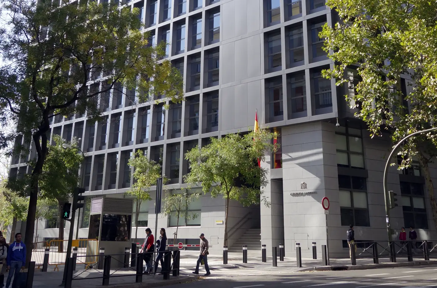 Fachada de la sede de la Audiencia Nacional en Madrid, un edificio moderno de ventanas rectangulares enmarcadas en gris, con la bandera de España y el escudo junto a la entrada, árboles y viandantes en la acera.