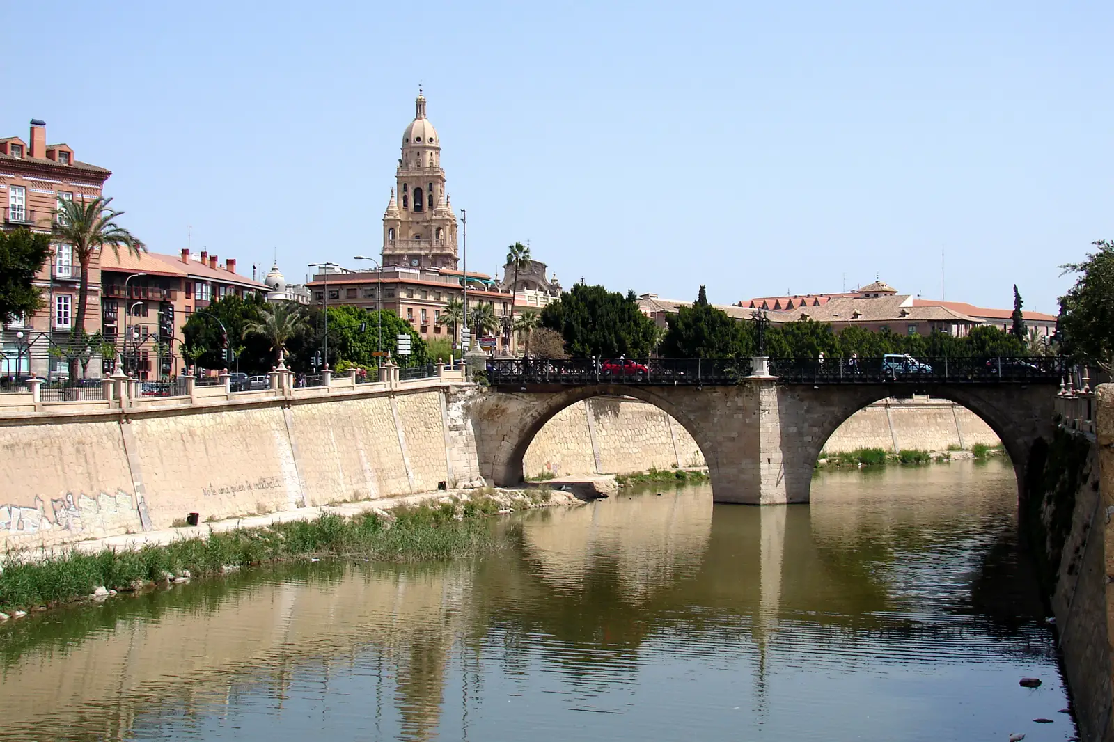 El río Segura a su paso por la ciudad de Murcia, encajonado entre altos muros de hormigón, con el agua muy baja y verdosa reflejando los dos arcos de piedra del puente de los Peligros; a la izquierda, casas de fachada rosa y palmeras del malecón y, al fondo, la torre barroca de la catedral bajo un cielo azul sin nubes.