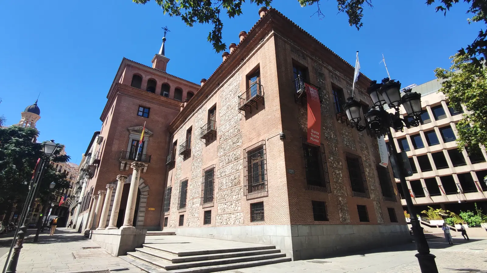 Fachada de ladrillo y mampostería de la casa de las Siete Chimeneas, un palacio del siglo XVI en Madrid, con una bandera de España sobre la portada principal y un cartel vertical granate colgado del muro.