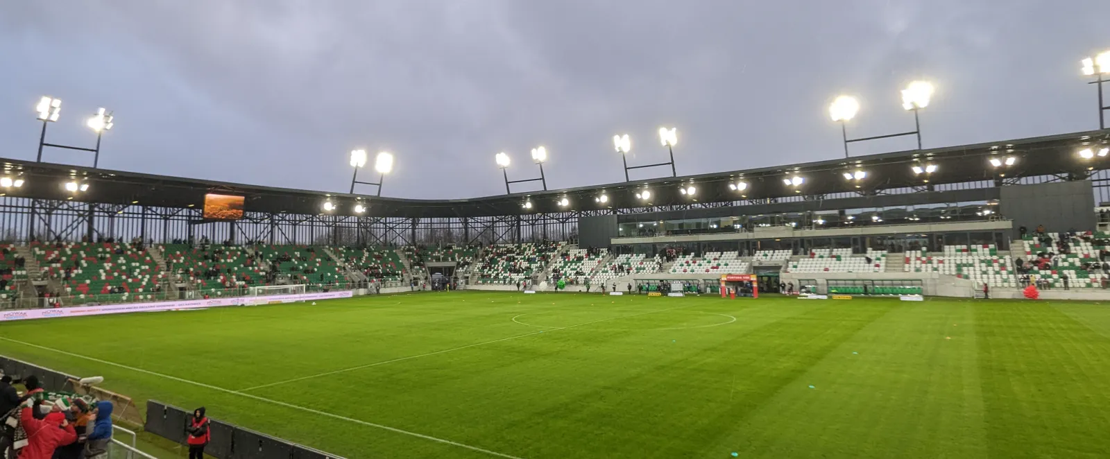 Interior del estadio del Zagłębiowski Park Sportowy de Sosnowiec al atardecer, con el césped iluminado por los focos y las gradas verdes y blancas parcialmente ocupadas.