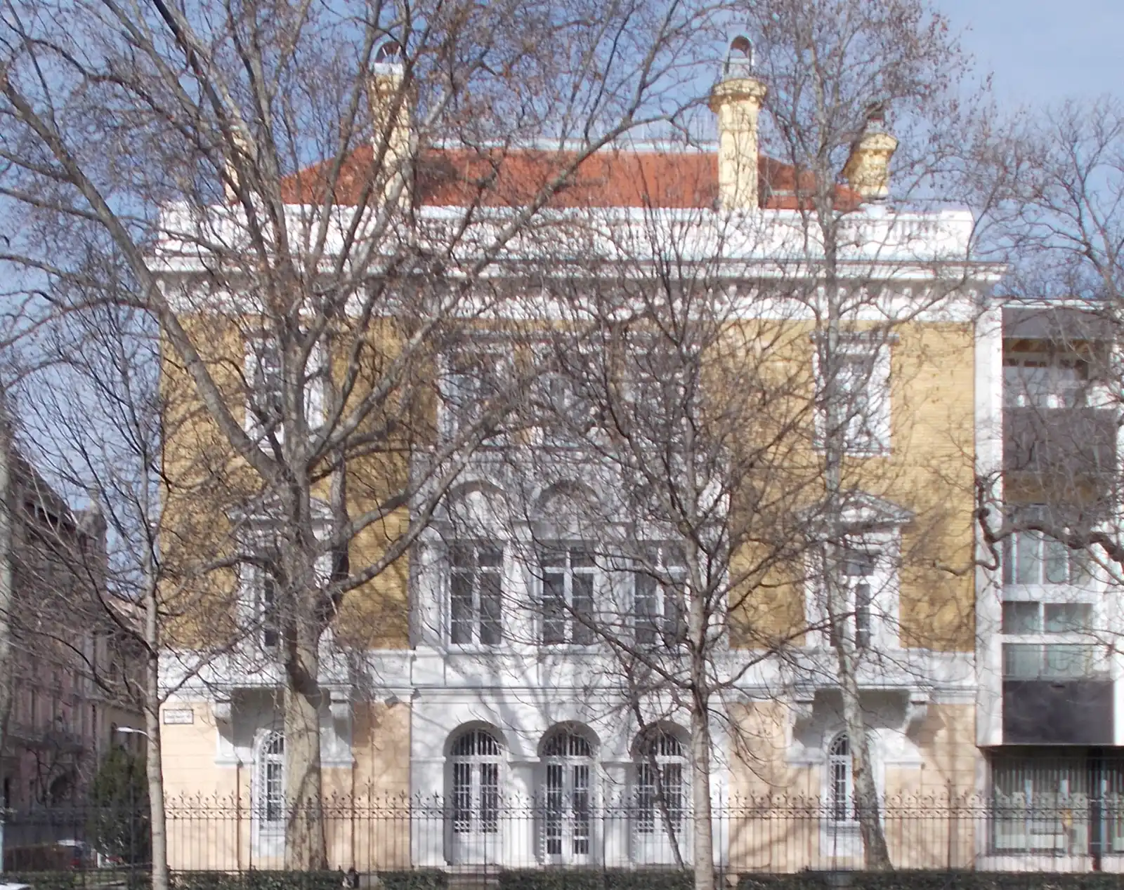 Palacete de dos plantas con fachada blanca y amarilla y tejado de tejas rojas, sede de la Embajada de Rusia en Budapest, visto tras una verja de hierro y varios árboles sin hojas