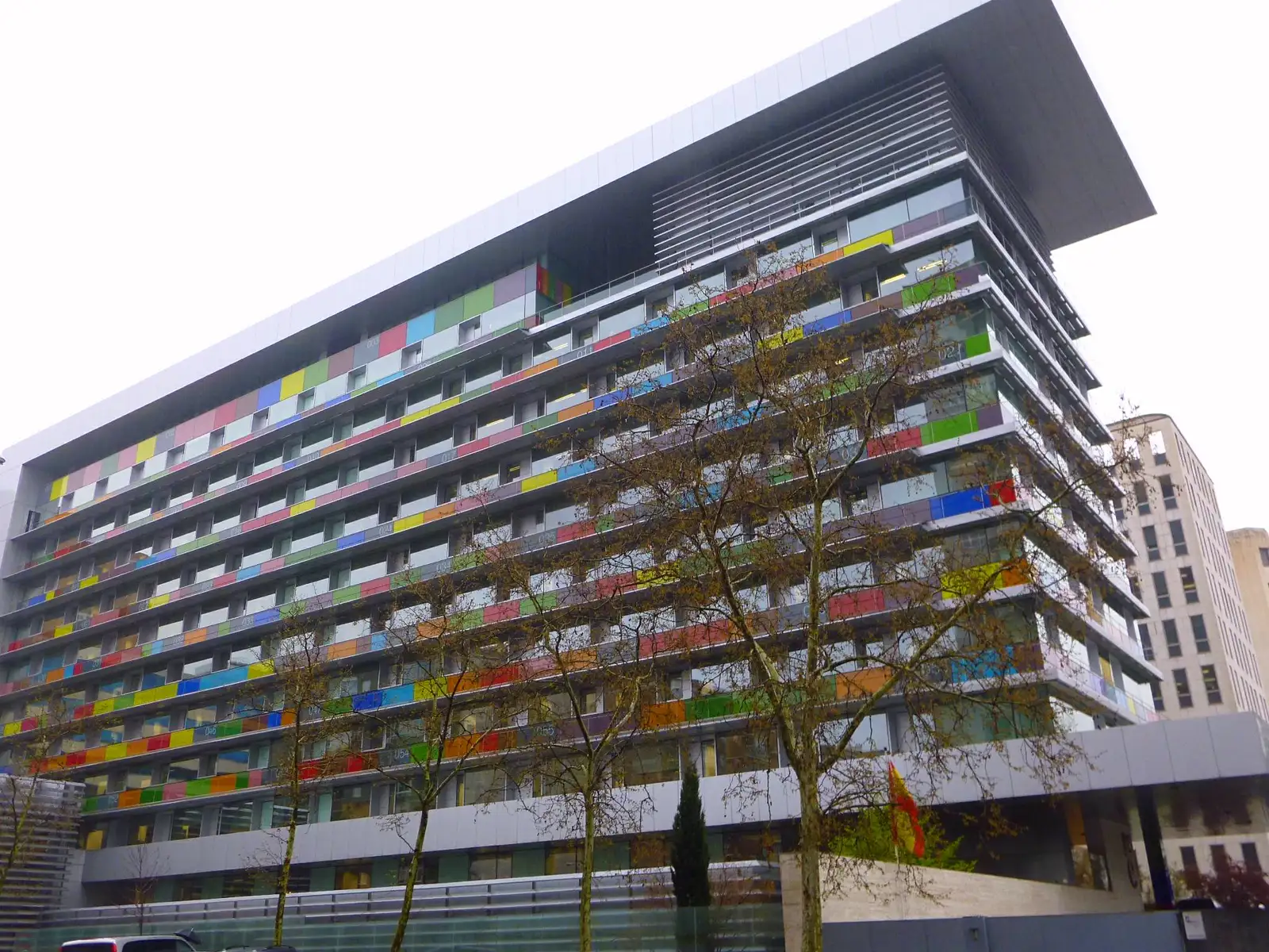 Fachada del edificio del Instituto Nacional de Estadística en Madrid, con paneles de colores entre las ventanas y árboles sin hojas delante