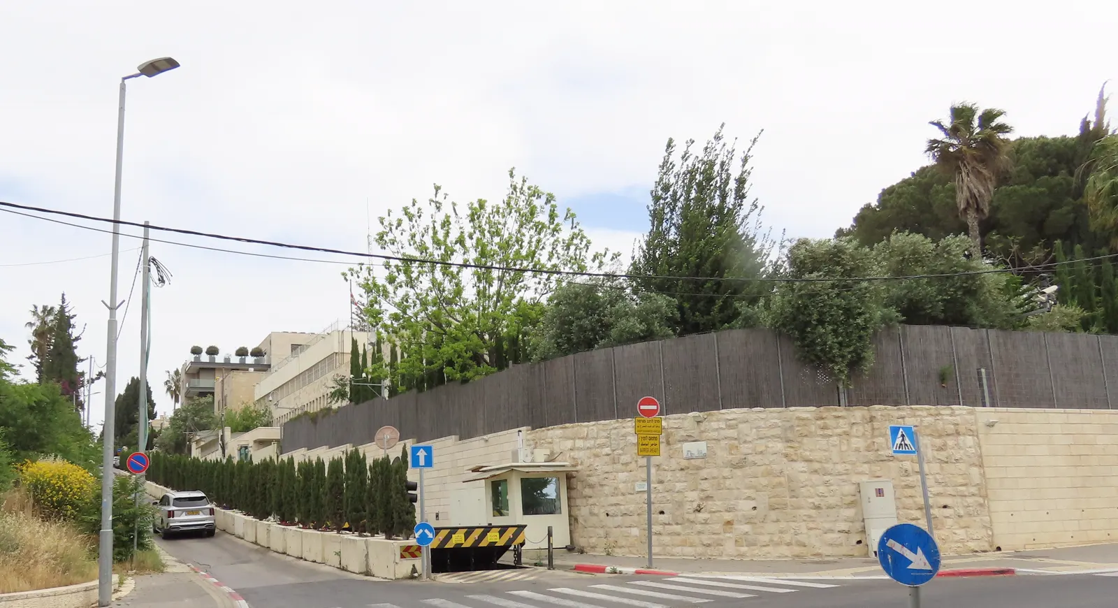 Recinto amurallado del consulado general británico en Jerusalén visto desde la calle Har haZeitim: muro de piedra, valla, garita de control con barrera en la entrada y, al fondo, el edificio de la misión con un mástil de bandera.