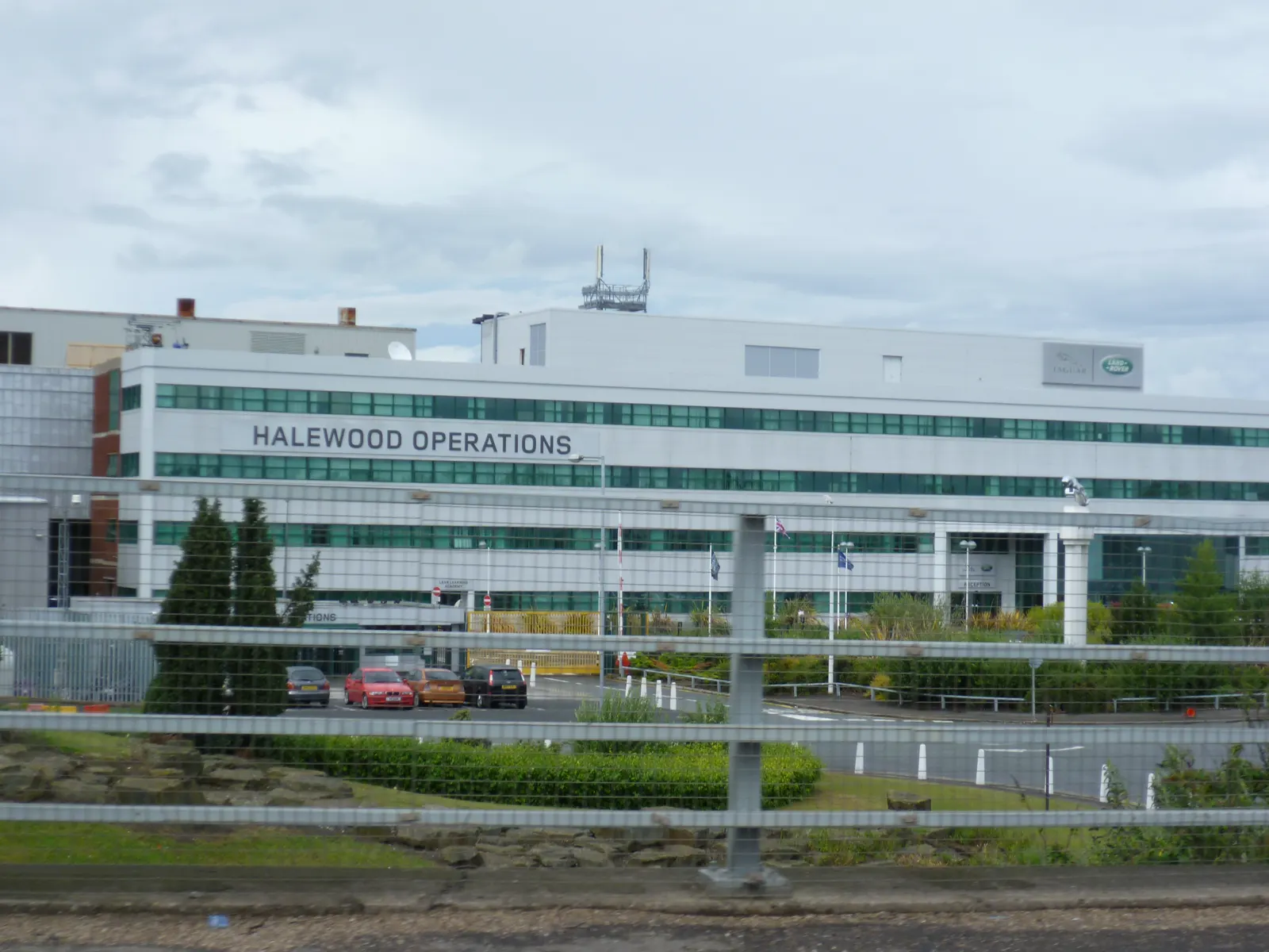 Edificio blanco y acristalado de varias plantas con el rótulo «Halewood Operations» y el logotipo de Land Rover en la fachada, visto desde el otro lado de una valla metálica, con coches aparcados en la entrada y cielo nublado.