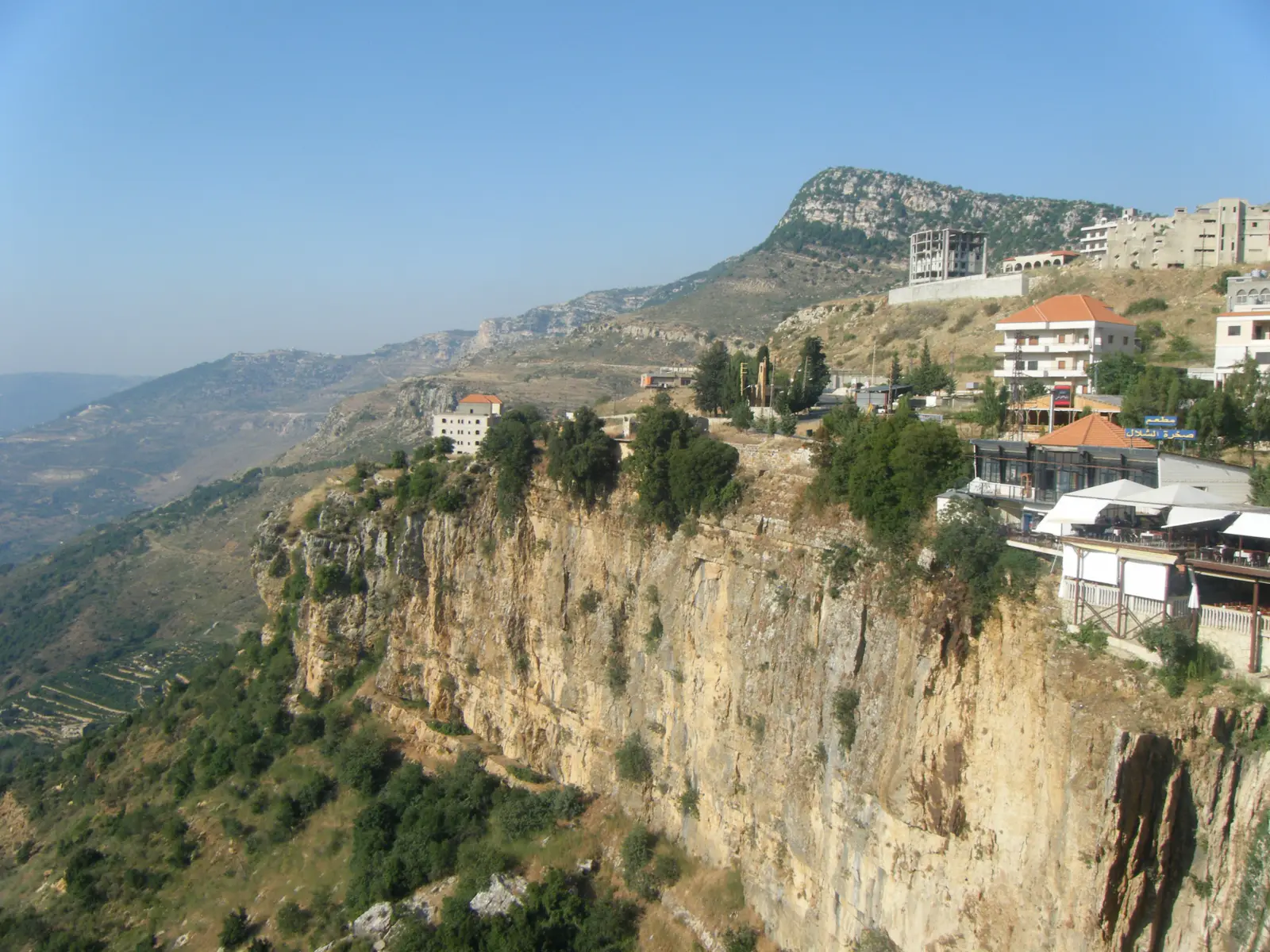Vista del pueblo de Jezzine, en el sur del Líbano, asomado al borde de un acantilado rocoso, con casas de tejado rojo, árboles y montañas peladas al fondo
