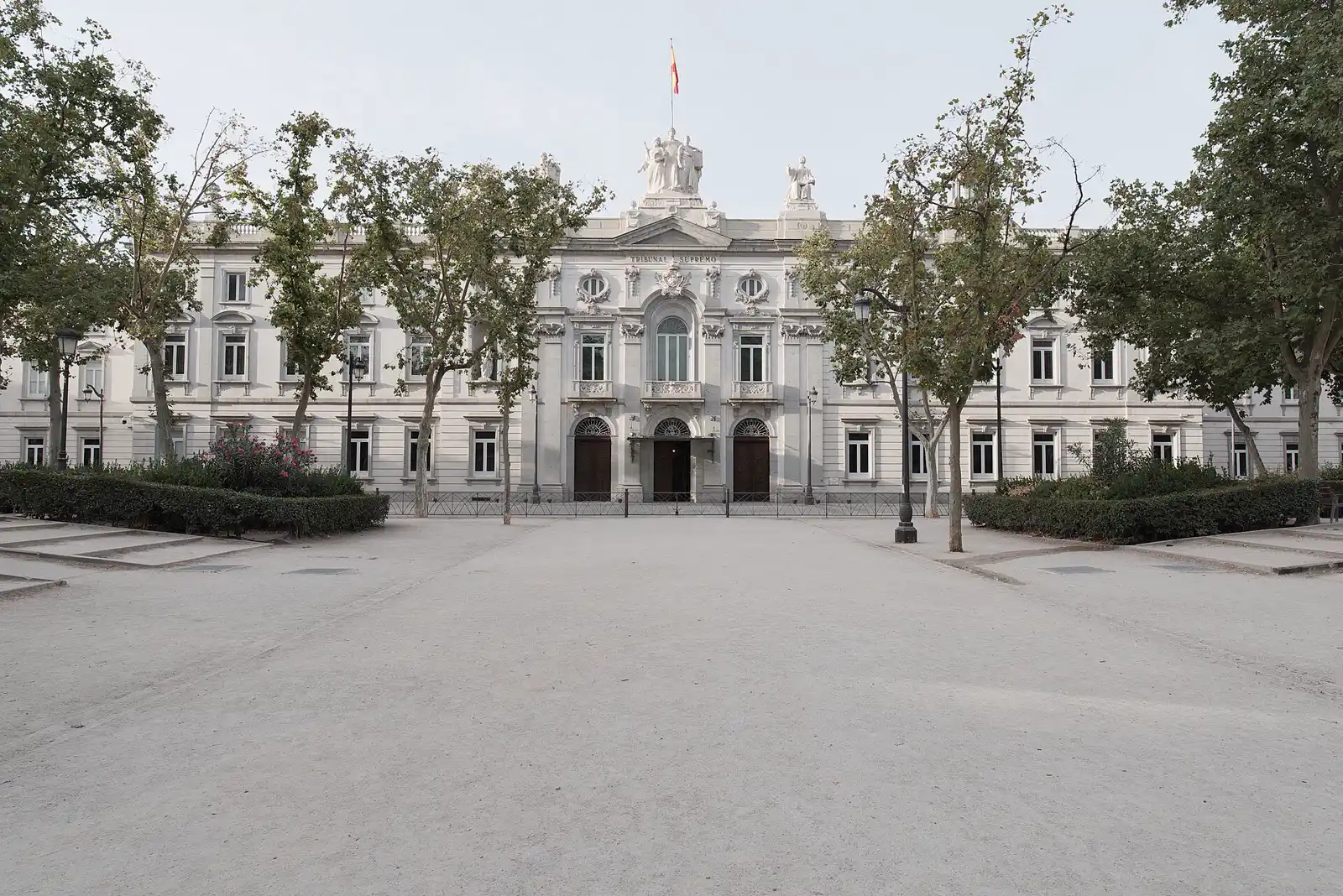 Fachada de piedra clara del Tribunal Supremo de Madrid vista de frente desde una explanada de albero vacía, con hileras de árboles a ambos lados, la bandera de España ondeando sobre el frontón y las tres puertas de acceso cerradas.