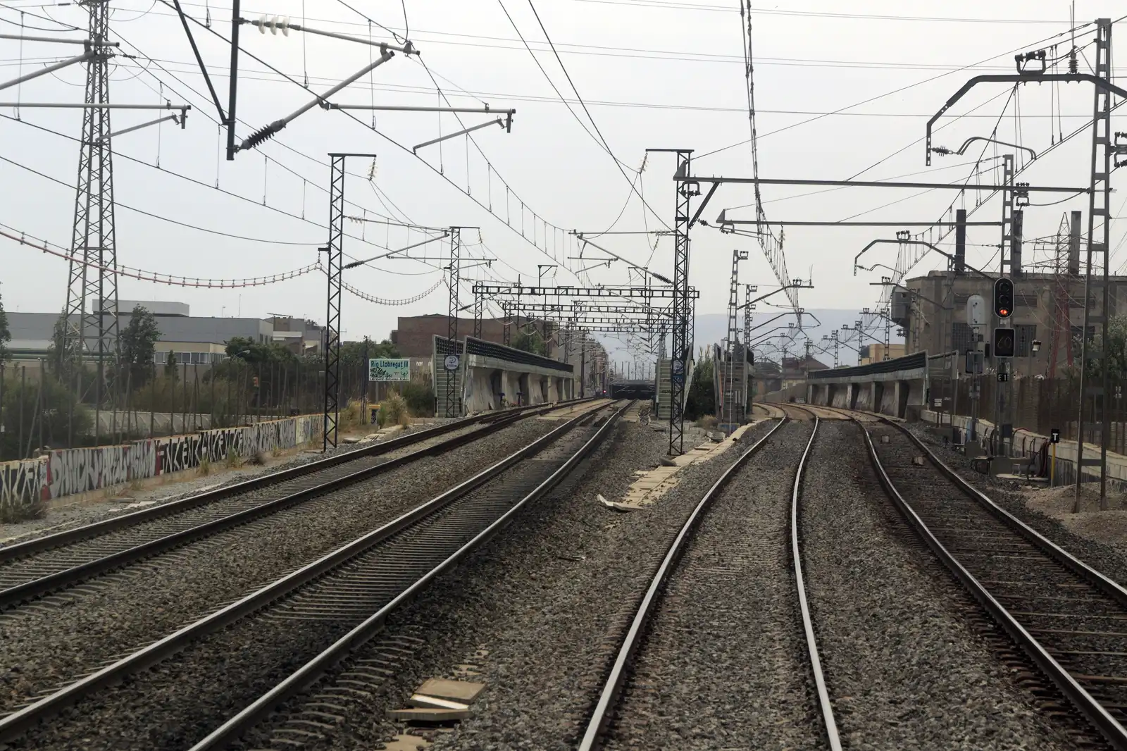 Haz de vías férreas que converge hacia el fondo entre balasto gris, bajo una maraña de postes, ménsulas y cables de catenaria; a la izquierda, una torre eléctrica de celosía y naves industriales; al fondo, los tableros de hormigón de dos puentes ferroviarios y una señal luminosa en rojo, con el cielo encapotado.