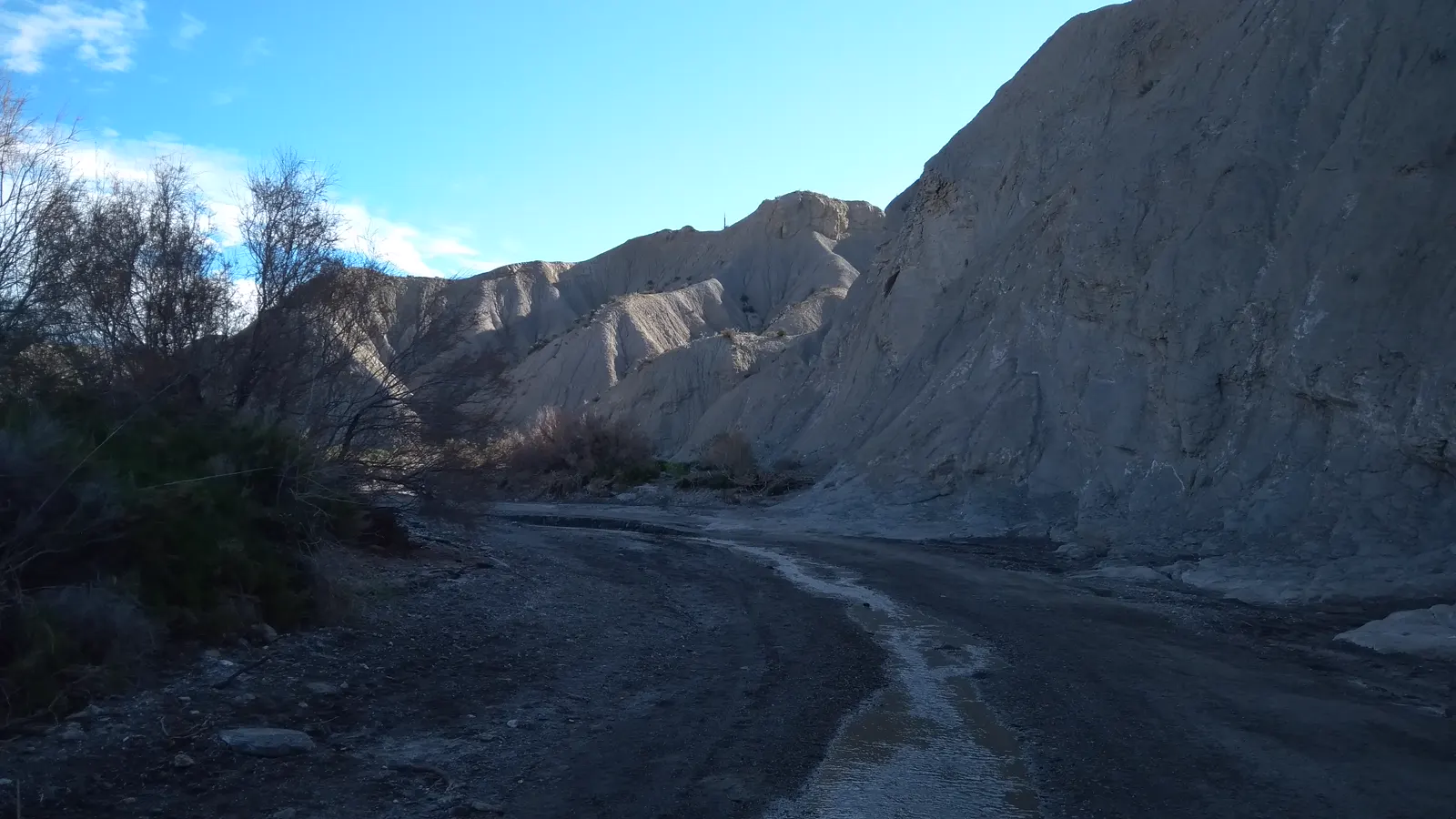 Cauce seco y pedregoso de una rambla que se curva entre altas paredes de tierra clara y erosionada; por el centro del lecho discurre un hilo de agua somera, con matorral y tarajes a la izquierda y cielo azul con algunas nubes.