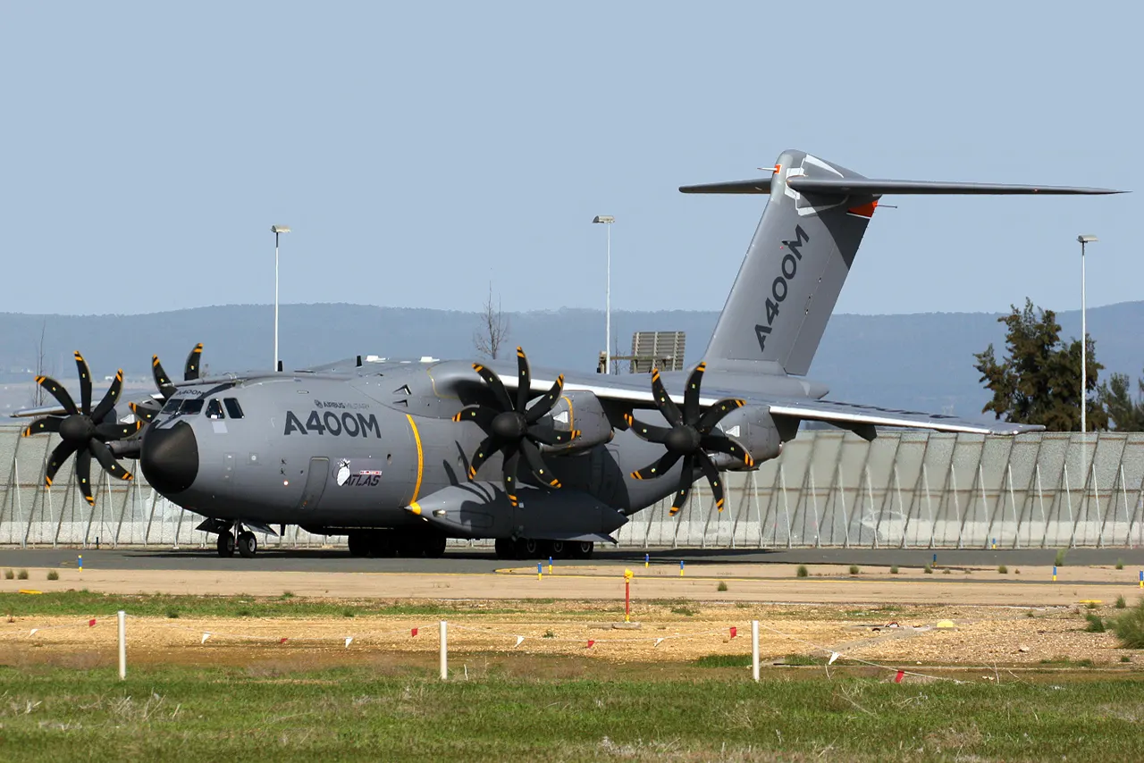 Avión de transporte militar Airbus A400M, pintado de gris y con cuatro motores de hélice, estacionado sobre una plataforma de asfalto en el aeropuerto de Sevilla. Al fondo, una valla perimetral y una línea de colinas bajo un cielo despejado.