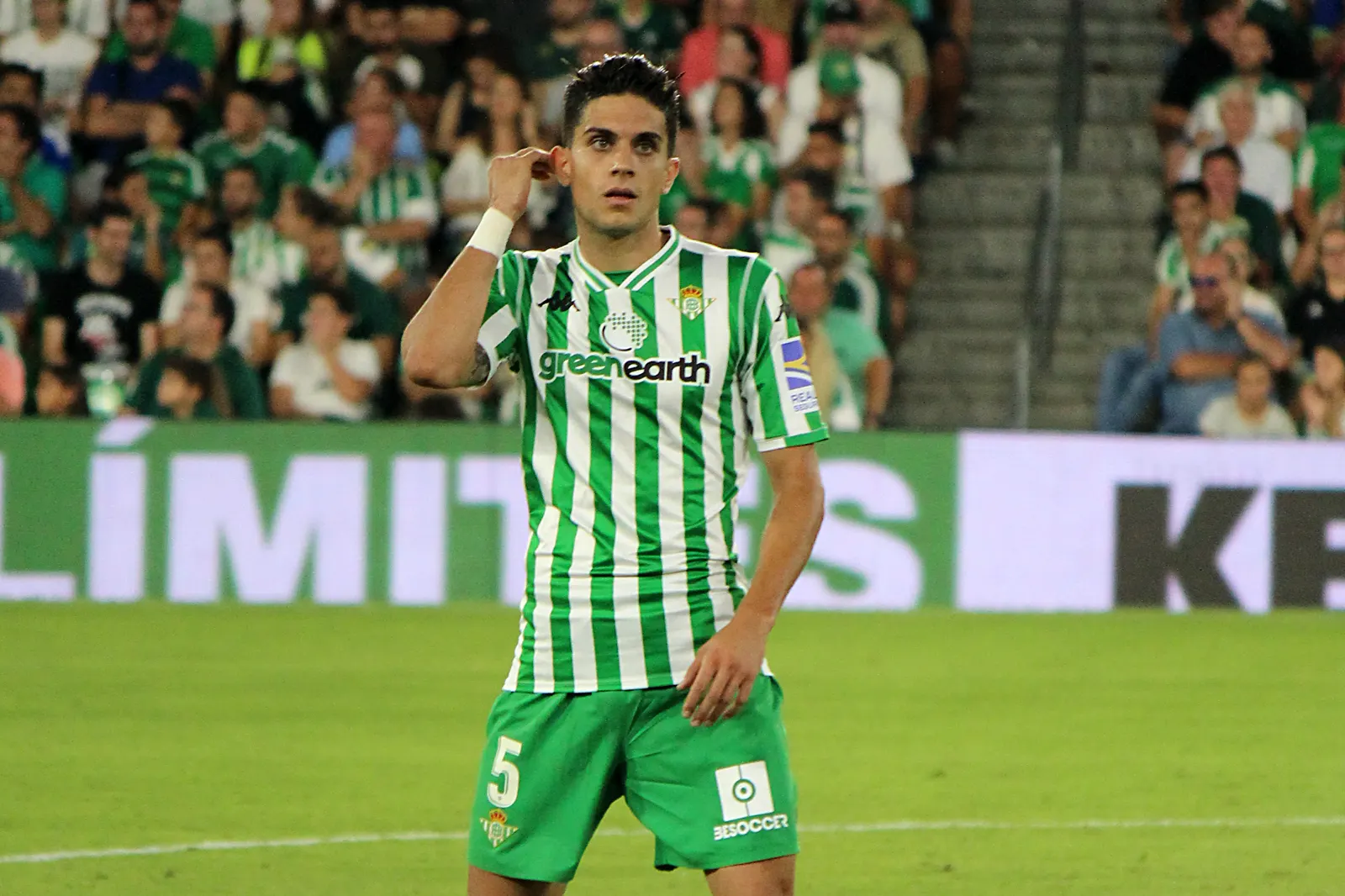 El futbolista Marc Bartra, con la camiseta a rayas verdiblancas del Real Betis y el dorsal 5, de pie sobre el césped durante un partido, con la grada llena al fondo