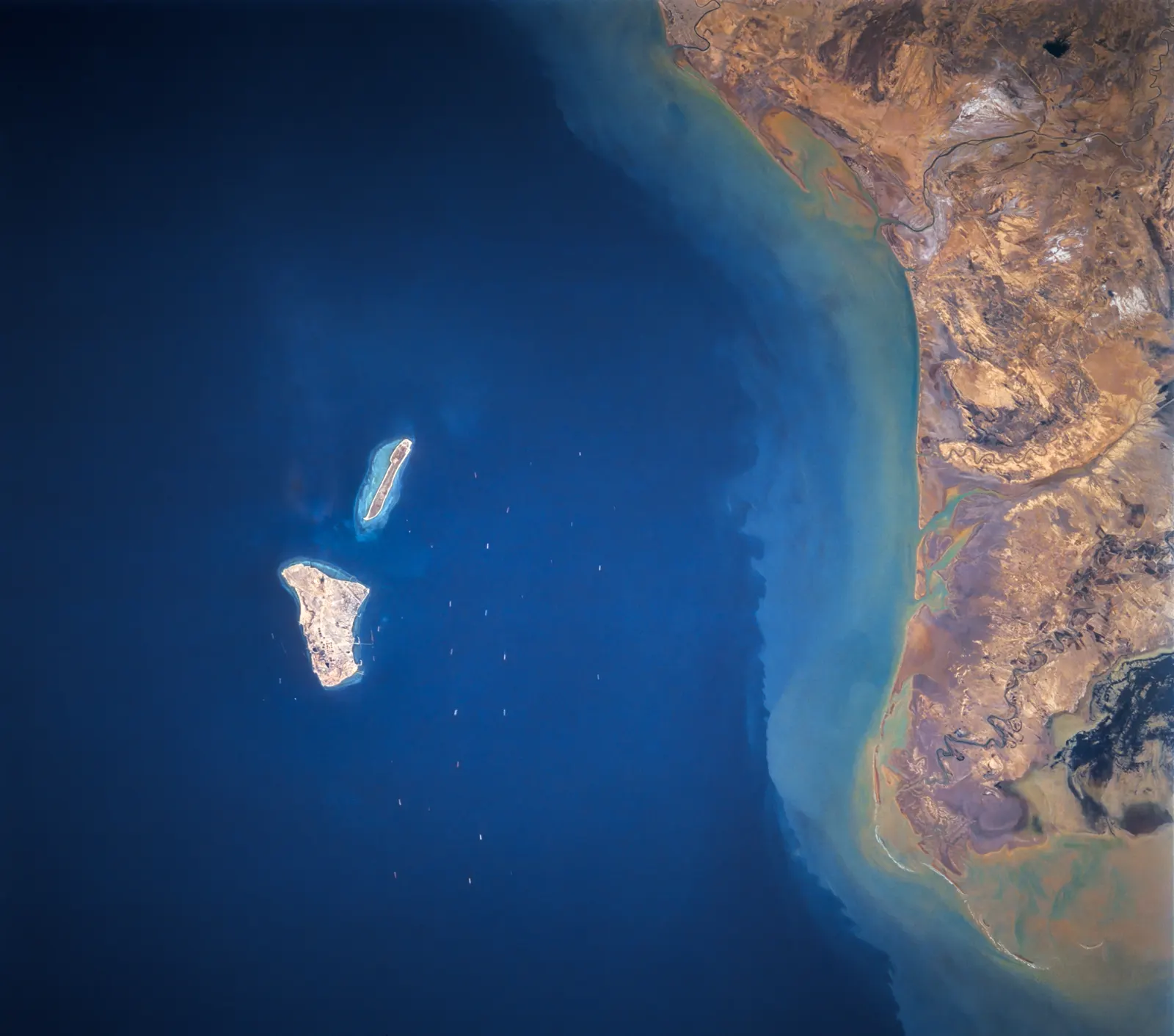 Fotografía desde el espacio de dos islas alargadas de tierra clara sobre un mar azul oscuro, frente a una costa árida y ocre surcada de barrancos. Alrededor de las islas se distinguen decenas de puntos blancos: buques fondeados.