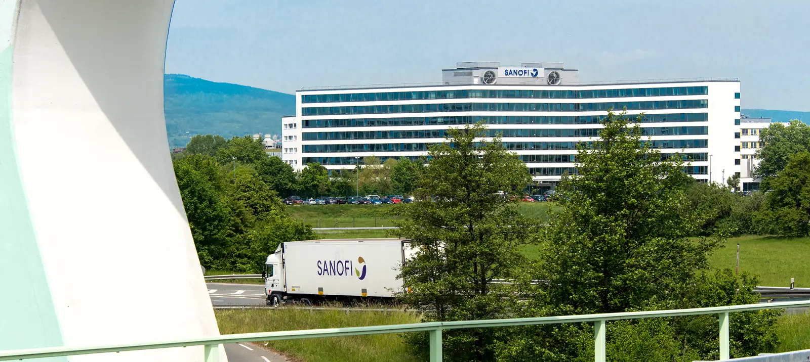 Edificio alargado y blanco de diez plantas con el logotipo azul de Sanofi en la azotea, rodeado de arboleda y con colinas boscosas al fondo; en primer plano, un camión con el rótulo «SANOFI» circula por una carretera junto a una pradera.