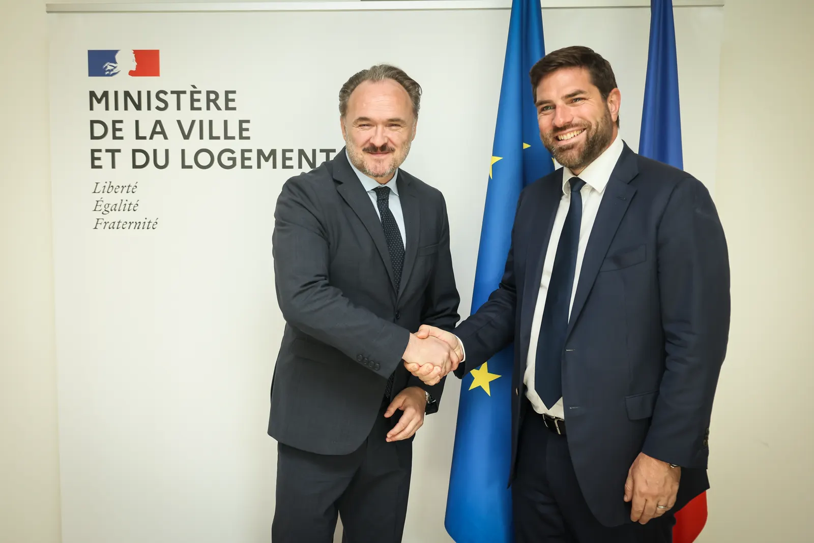 Dan Jørgensen, a la izquierda, con barba entrecana y traje oscuro, estrecha la mano de Vincent Jeanbrun ante un panel del Ministerio de la Ciudad y la Vivienda francés, con las banderas europea y francesa al fondo.