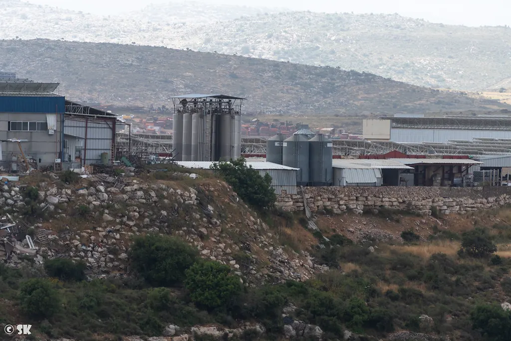 Naves industriales de chapa, silos metálicos y contenedores apilados en una ladera pedregosa, con colinas áridas al fondo.