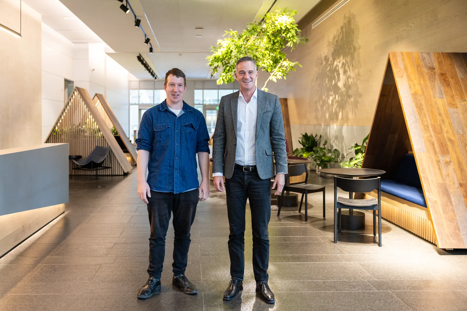Vestíbulo de las oficinas de Anthropic en San Francisco: dos hombres de pie sobre un suelo de piedra, entre alcobas de madera claras, sillones bajos, plantas y un árbol interior.