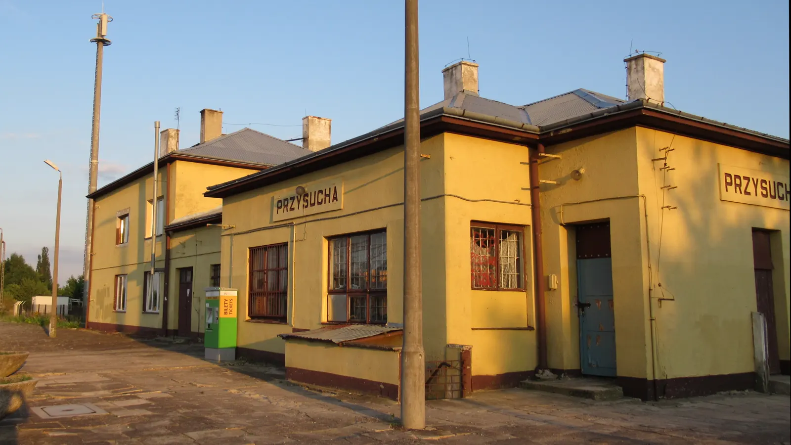 Edificio bajo de la estación ferroviaria de Przysucha, de fachada amarilla y con el rótulo PRZYSUCHA en letras rojas, junto a una máquina expendedora de billetes, al atardecer.