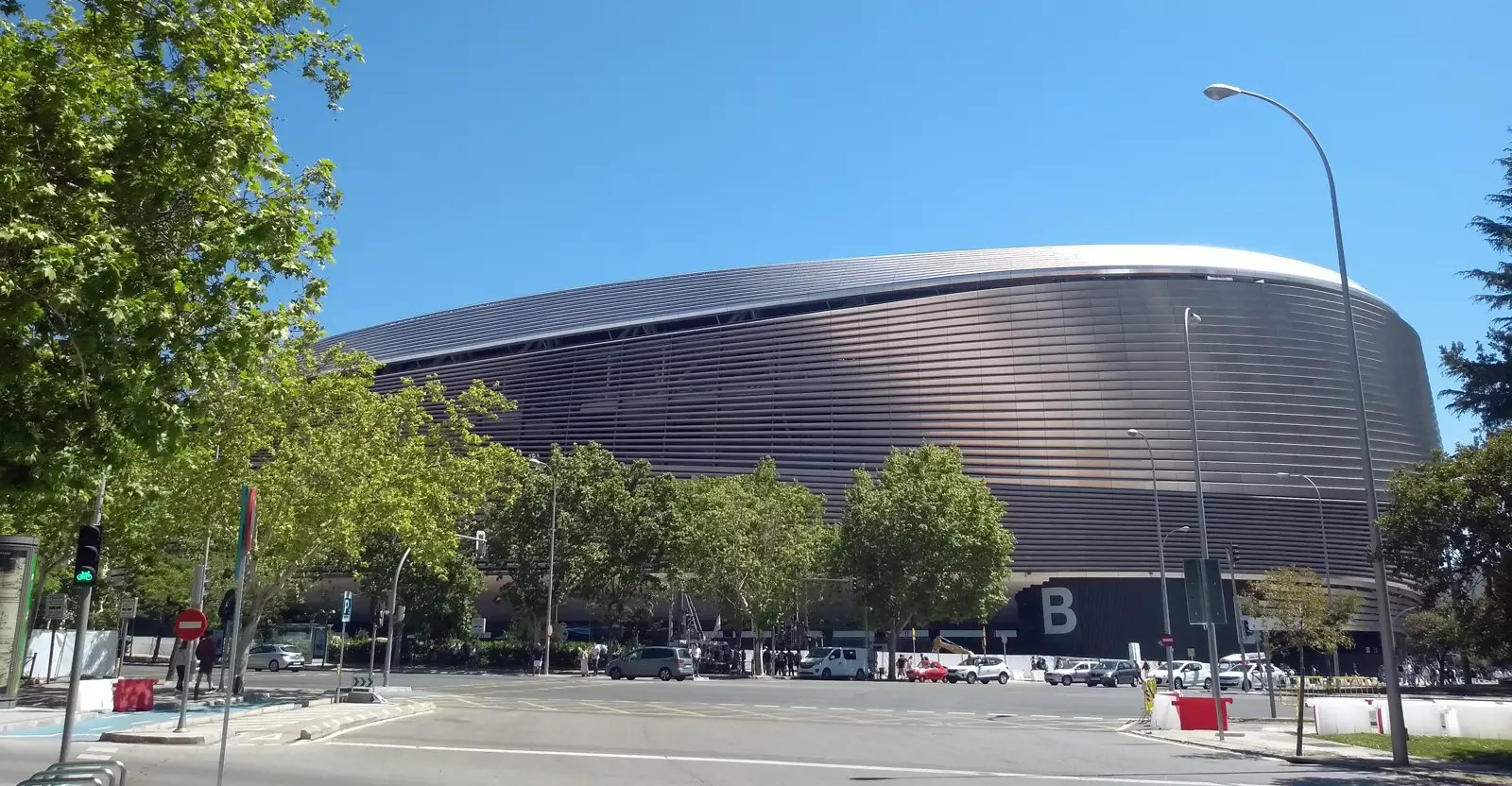 Fachada oeste del estadio Santiago Bernabéu, de líneas curvas y revestimiento metálico horizontal, vista desde la calle un día soleado, con árboles y tráfico en primer plano