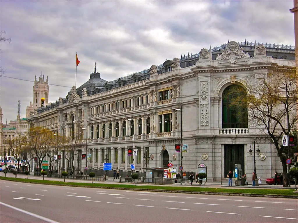 Fachada norte del Banco de España en la calle de Alcalá de Madrid, un edificio de piedra clara con arcos, columnas y relieves, con una bandera española ondeando sobre la cubierta, algunos peatones en la acera y la calzada vacía en primer plano bajo un cielo nublado.
