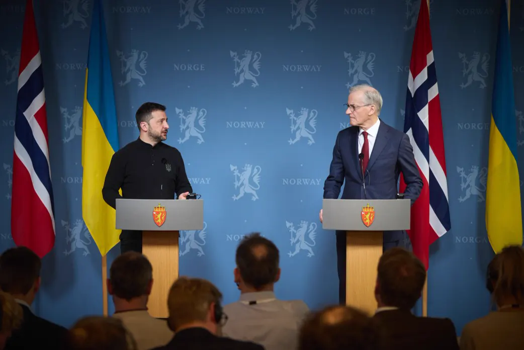 Volodímir Zelenski y Jonas Gahr Støre, de pie tras sendos atriles con el escudo del león noruego, se miran durante una rueda de prensa; al fondo, un panel azul con la palabra NORWAY y banderas de Noruega y Ucrania; en primer plano, las nucas del público.