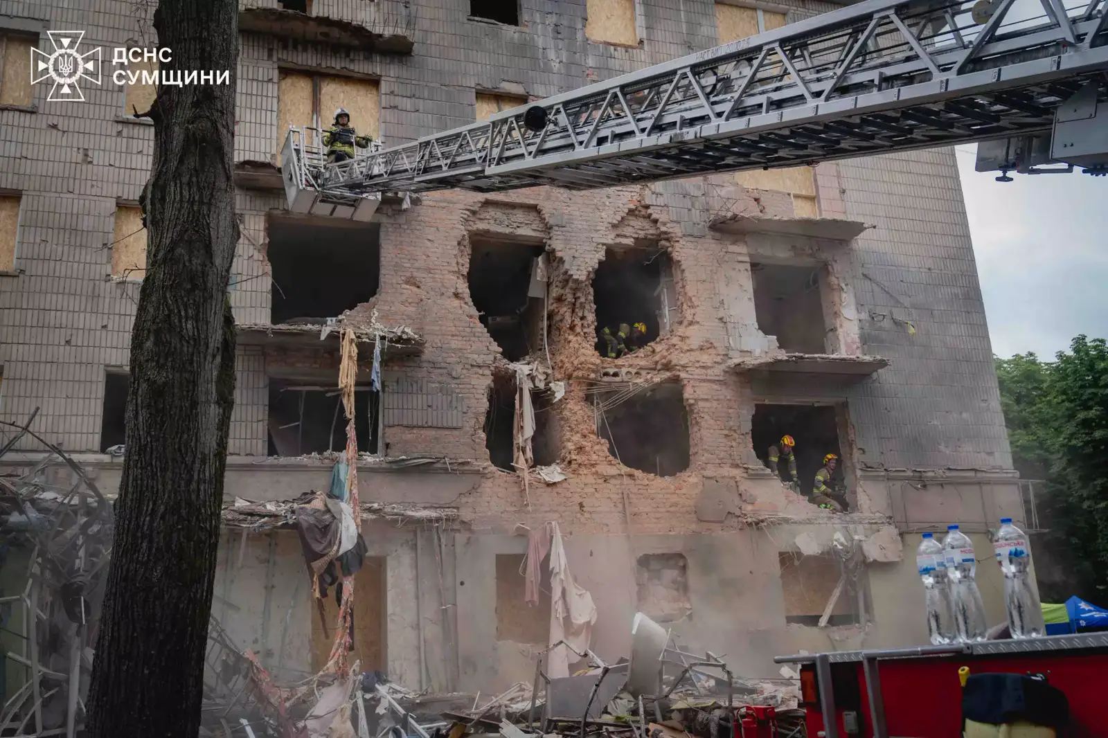Bomberos con casco trabajan en la fachada reventada de un edificio de Sumi, con boquetes de varios metros en el muro de ladrillo, escombros en el suelo y un camión escalera desplegado hasta una de las plantas.