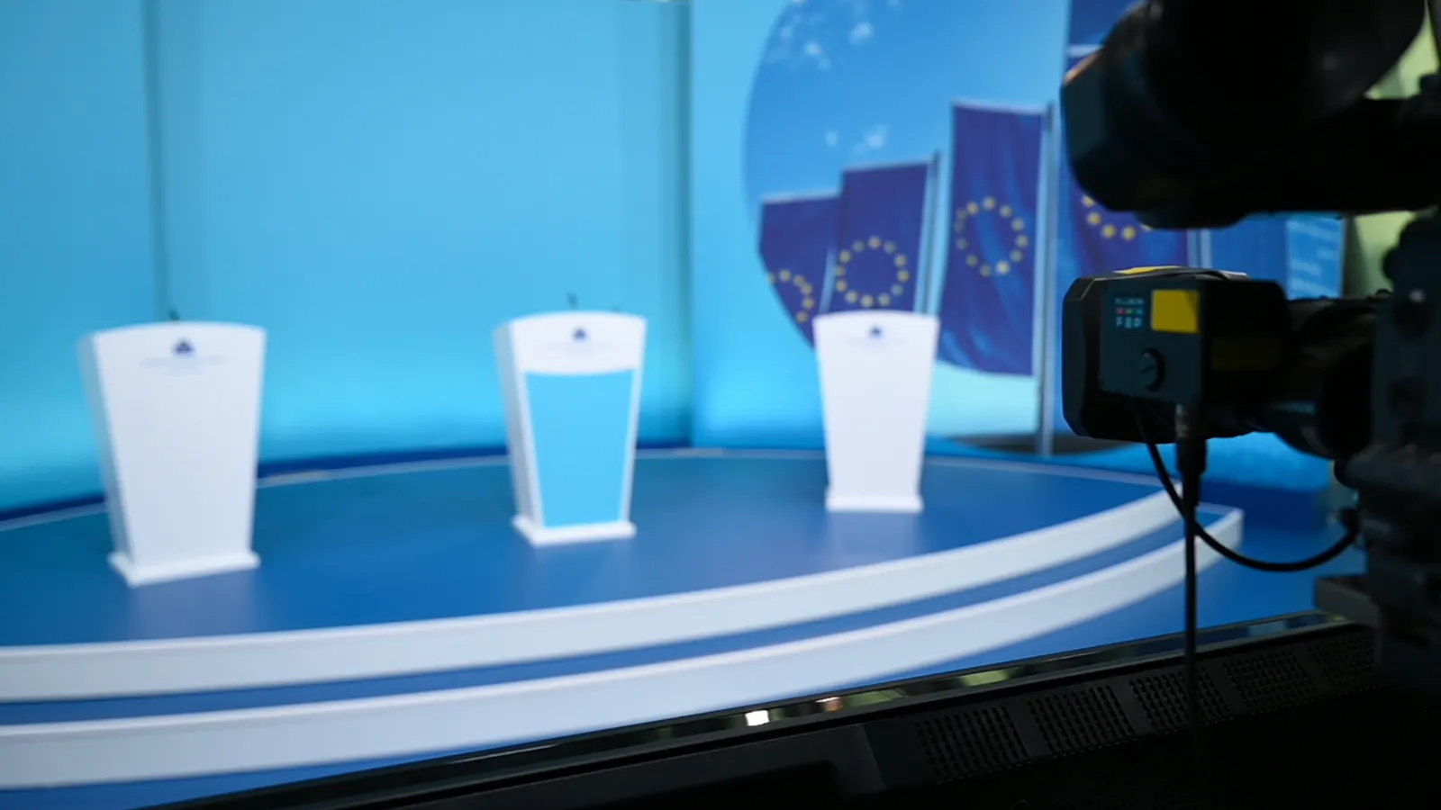 Sala de rueda de prensa del Banco Central Europeo, con tres atriles blancos sobre una tarima azul, banderas de la Unión Europea al fondo y una cámara de televisión en primer plano.