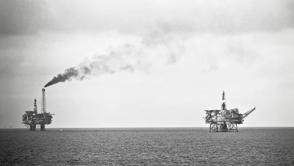 Fotografía en blanco y negro de dos plataformas petrolíferas sobre un mar en calma bajo un cielo encapotado. La de la izquierda emite una columna de humo negro desde su antorcha; la de la derecha, con una grúa, aparece recortada contra el horizonte.
