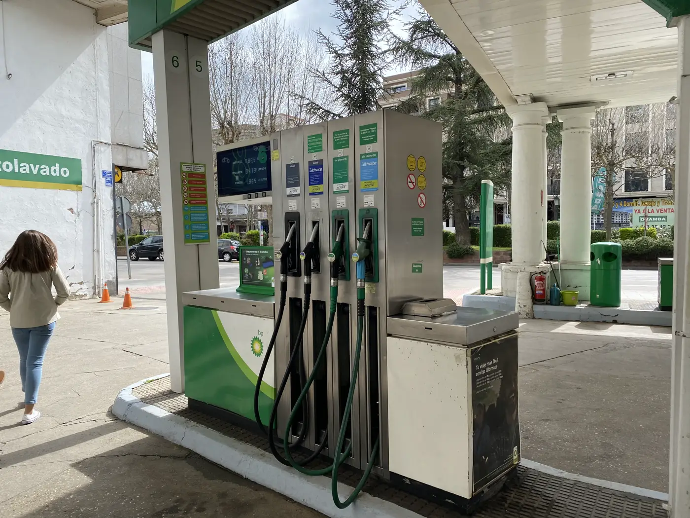 Isla de surtidores de una gasolinera BP bajo su marquesina, con cuatro mangueras verdes y negras colgadas y una pantalla de precios encendida sobre el dispensador. A la izquierda, una mujer de espaldas se aleja caminando hacia el túnel de lavado; al fondo, árboles y edificios de una calle urbana.