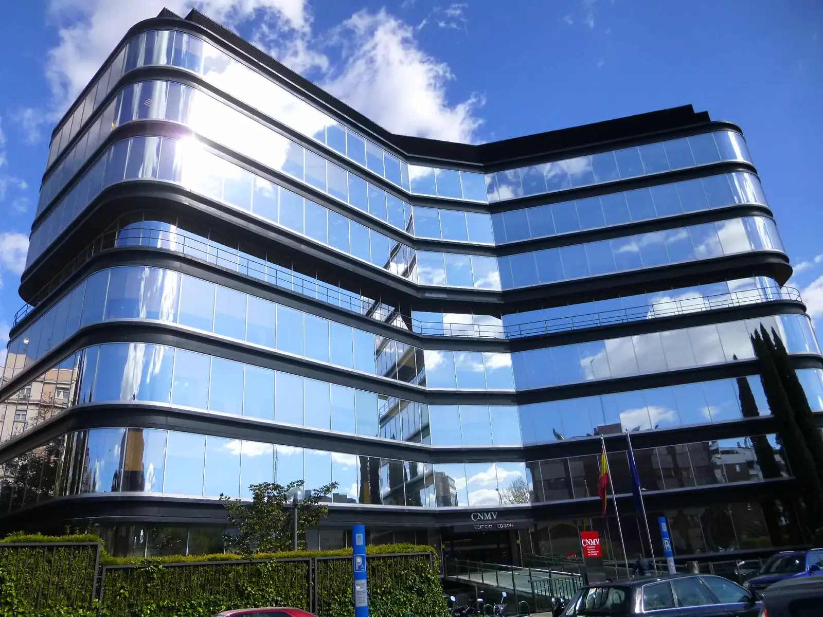 Edificio de oficinas de siete plantas con fachada curva de cristal espejado que refleja el cielo; sobre la entrada, el rótulo CNMV y, a la derecha, un cartel rojo del organismo junto a las banderas de España y la Unión Europea.