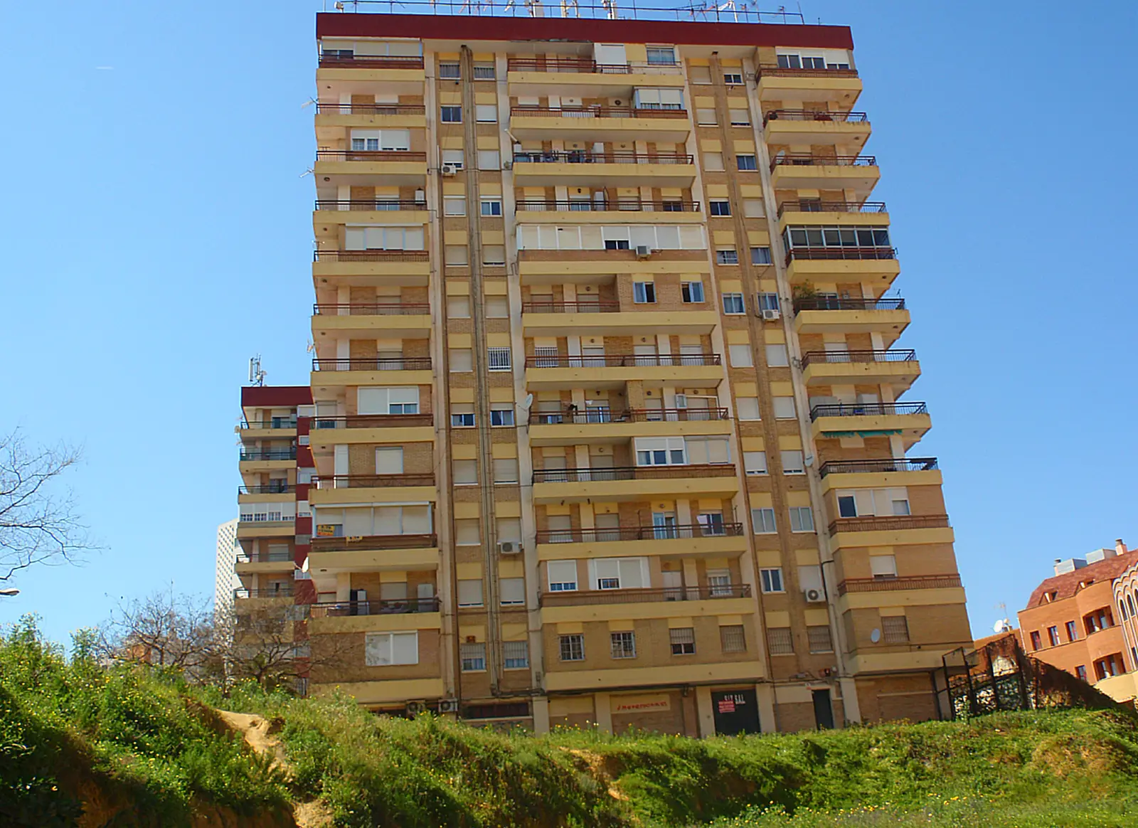 Torre de viviendas de doce alturas en Huelva, de ladrillo y balcones corridos, vista desde abajo contra un cielo azul, con un talud de vegetación en primer plano.