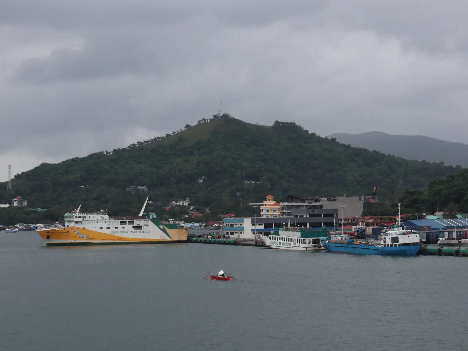 Vista del puerto de Coron, en Palawan, con dos ferris de pasajeros y un carguero atracados junto al muelle y el monte Tapyas al fondo, bajo un cielo nublado