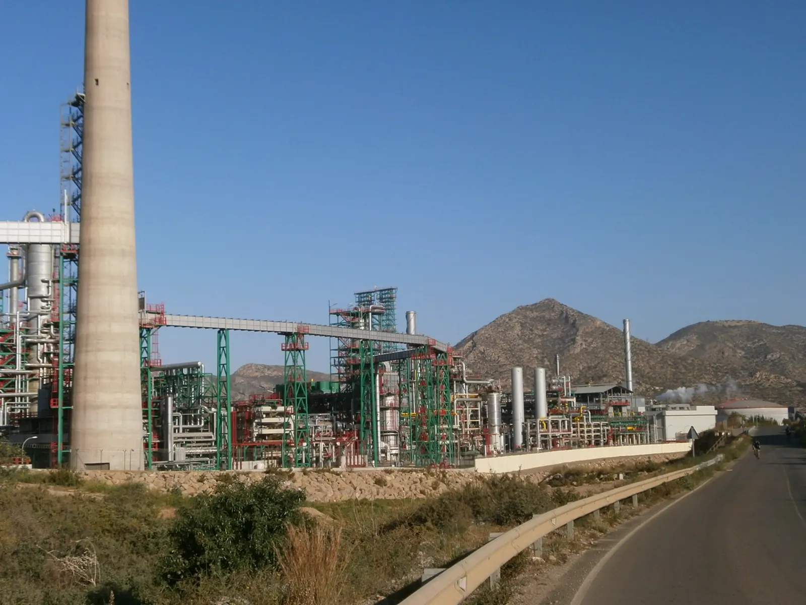 Instalaciones de una refinería junto a una carretera: una chimenea de hormigón muy alta, torres de destilación, pasarelas y estructuras metálicas verdes y rojas, con cerros áridos y cielo despejado al fondo.