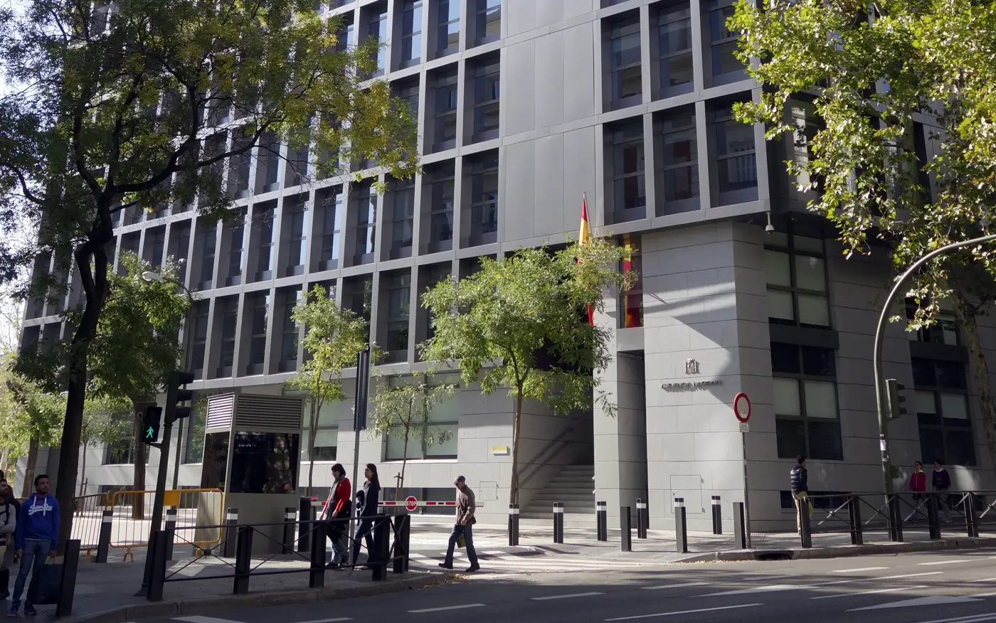 Fachada de la sede de la Audiencia Nacional en Madrid: un edificio moderno de cristal y hormigón gris con la bandera de España junto a la entrada, el escudo y el rótulo «Audiencia Nacional» sobre el zócalo de piedra, con árboles y viandantes en la acera.